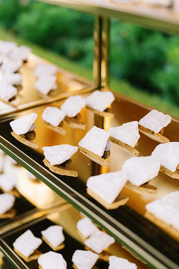 Wedding place cards arranged on a gold mirrored escort card display with white calligraphy lettering against lush garden greenery backdrop