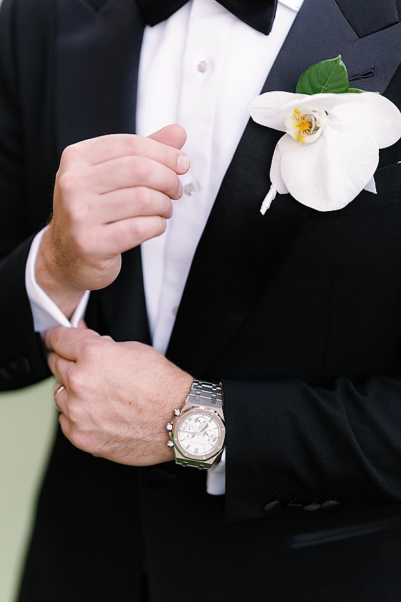 Groom boutonniere with a white orchid boutonniere pinned to a black tuxedo jacket, bow tie and wristwatch visible against blurred greenery