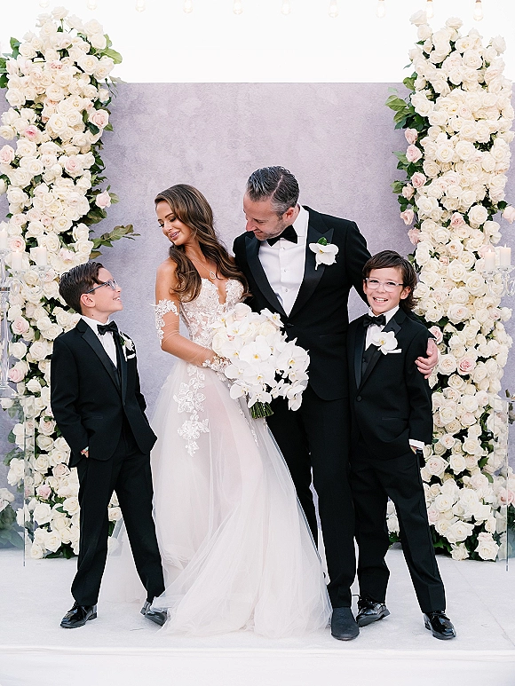 Family wedding portrait of bride and groom with kids, bride holding a white orchid bouquet beside boys in tuxedos on a gray backdrop