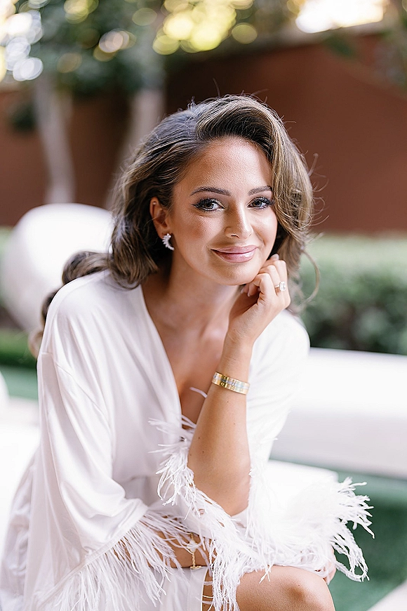 Bridal portrait of a getting ready bride in a white feather-trim robe, seated on an outdoor patio lounge chair with greenery and sunlit trees