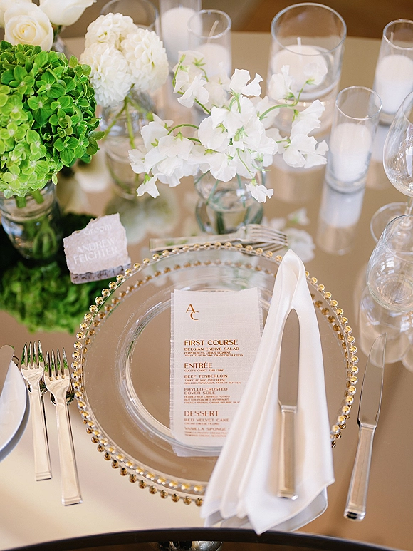 Reception tablescape with wedding place setting featuring a clear beaded charger plate, menu card, white napkin, silver flatware, and floating candle centerpiece
