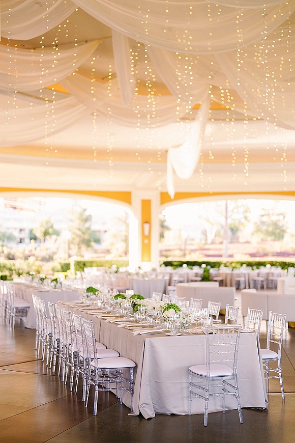 Reception tablescape with a white wedding tablescape featuring greenery centerpieces and string lights under draped fabric on a covered patio with arches