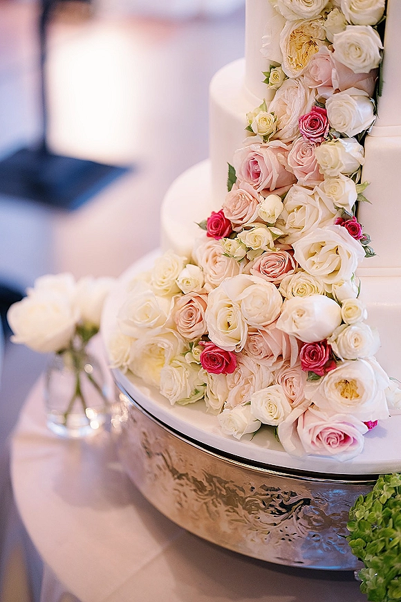 Wedding cake with a rose cascade of blush and ivory roses over buttercream tiers on a stand atop white linen in a blurred reception space