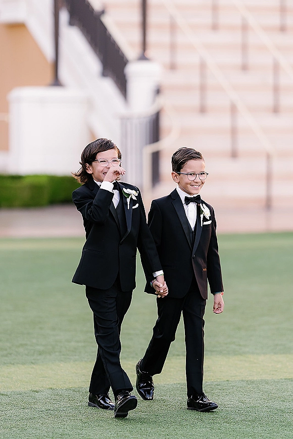 Ring bearer portrait of two ring bearers holding hands in black tuxedos with bow ties, one in eyeglasses, on a lawn by steps