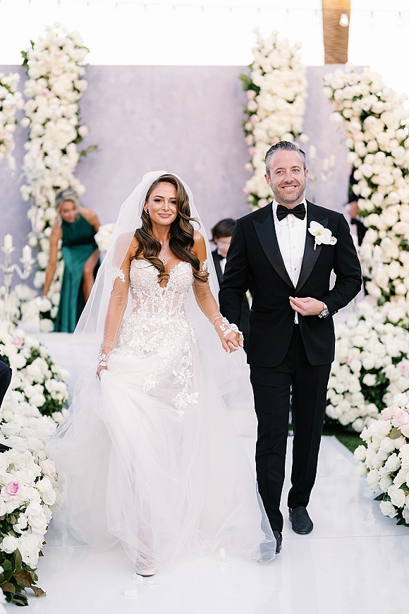 Recessional moment as bride and groom walking down aisle holding hands, her cathedral veil and lace sleeves flowing past white rose florals