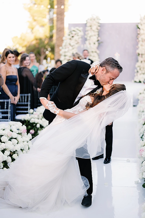 Wedding kiss as groom dips bride in lace dress and veil, guests watching along a flower-lined outdoor aisle with trees behind
