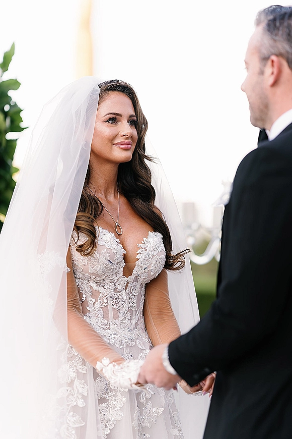 Ceremony moment as bride and groom holding hands, bride gazing at groom in lace dress with veil, outdoors under bright sky
