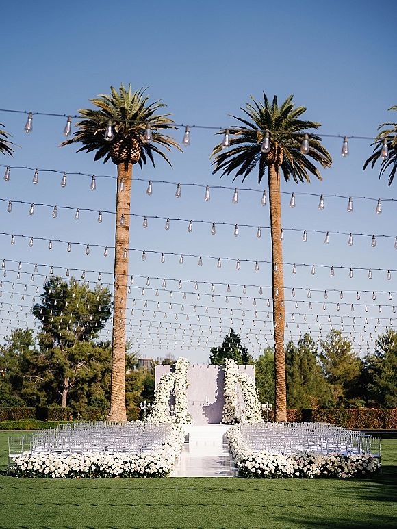 Ceremony setup for an outdoor wedding ceremony with clear acrylic chairs, white floral altar, and flower-lined aisle on a palm-lined lawn
