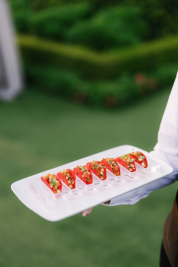 Wedding appetizers on a white serving platter, held by a gloved server with bite-size hors d'oeuvres on a green lawn by hedges