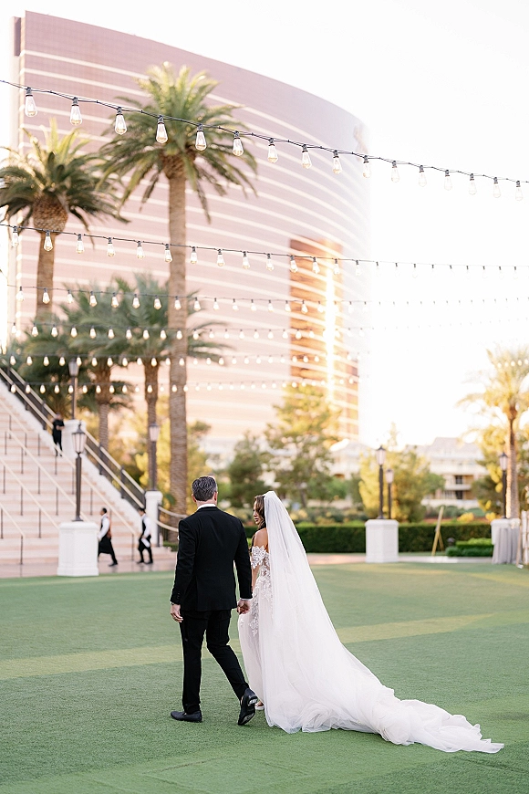 Couple portrait of bride and groom walking hand in hand, lace dress and veil train flowing under string lights by palm trees near a modern hotel lawn
