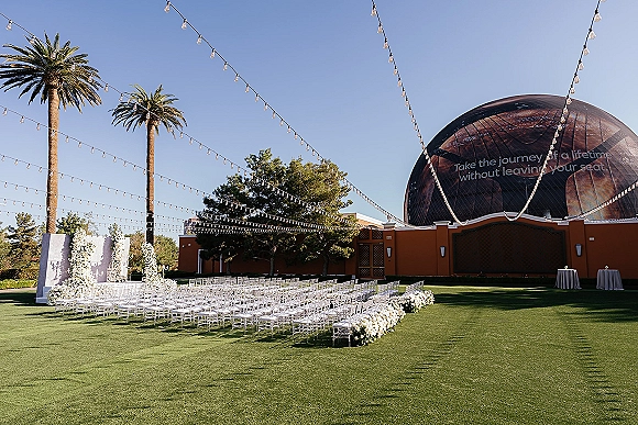 Outdoor ceremony setup with string lights and white floral backdrop, clear chairs and aisle flowers on a green lawn with palm trees