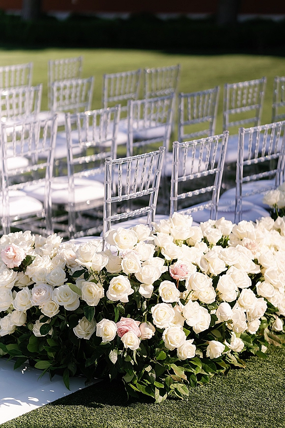 Ceremony aisle decor with a floral lined aisle of white and blush roses and greenery garland bordering a white runner on a lawn