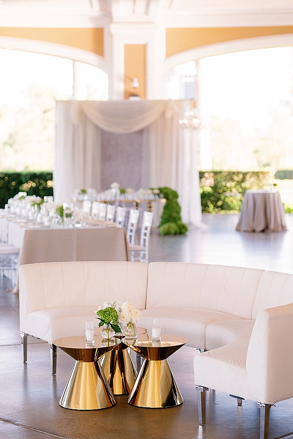 Wedding lounge seating with a white curved sofa, gold coffee tables, floral arrangements and glass votive candles in an indoor reception space
