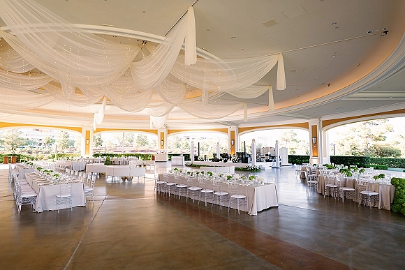 Reception setup with wedding reception layout under ceiling drape and string lights, white linens, clear chairs, and green floral centerpieces in a ballroom