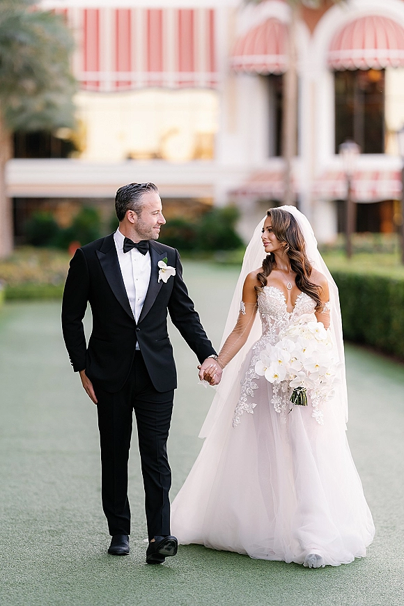 Couple portrait of bride and groom holding hands, her lace gown and veil flowing as they walk past hedges and striped awnings