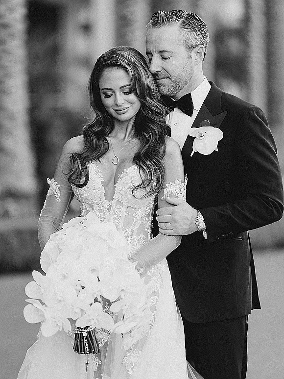 Wedding couple portrait of bride and groom embrace, forehead touch by columned outdoor walkway, bride in lace gown holding orchid bouquet