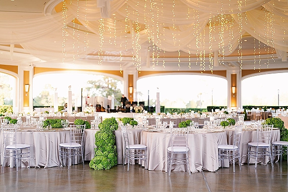 Reception tablescape with ivory linens, round tables, clear Chiavari chairs, and green centerpieces under string lights in an open-air pavilion