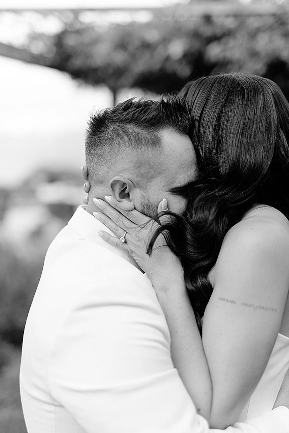 Wedding kiss portrait in black and white, close-up of couple embracing as bride cups groom’s face, ring visible against blurred greenery