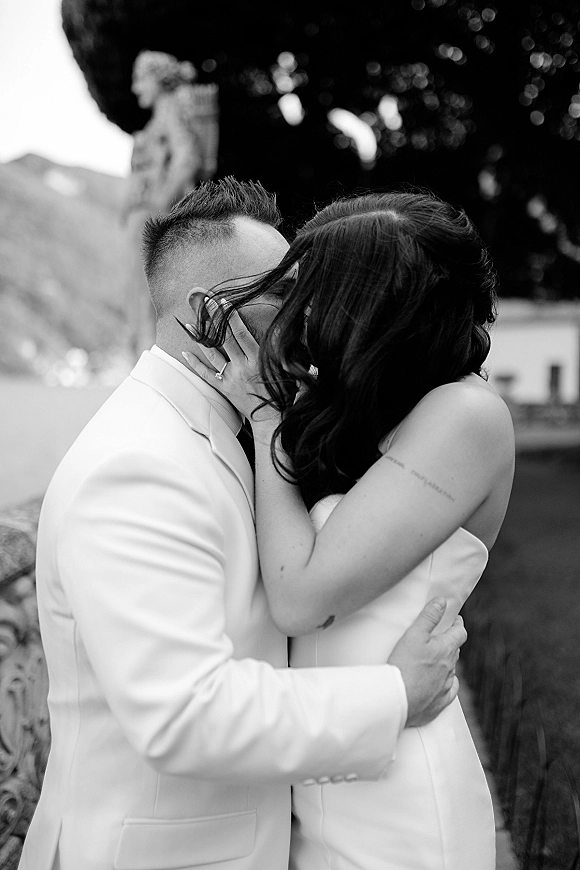 Wedding kiss portrait in black and white as bride cups groom’s face, ring visible, in a garden with trees and a stone statue backdrop