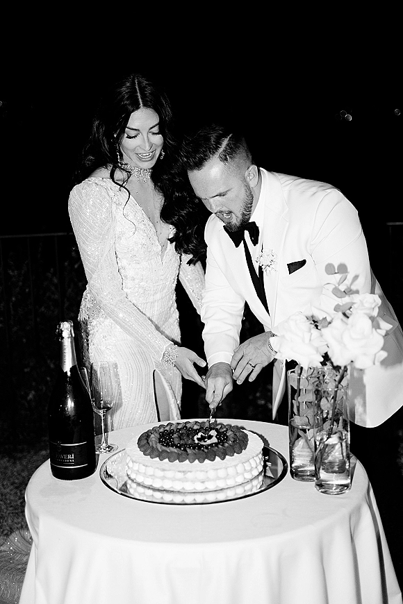 Wedding cake cutting as bride and groom slice a fruit-topped cake on a small round table with champagne under the night sky