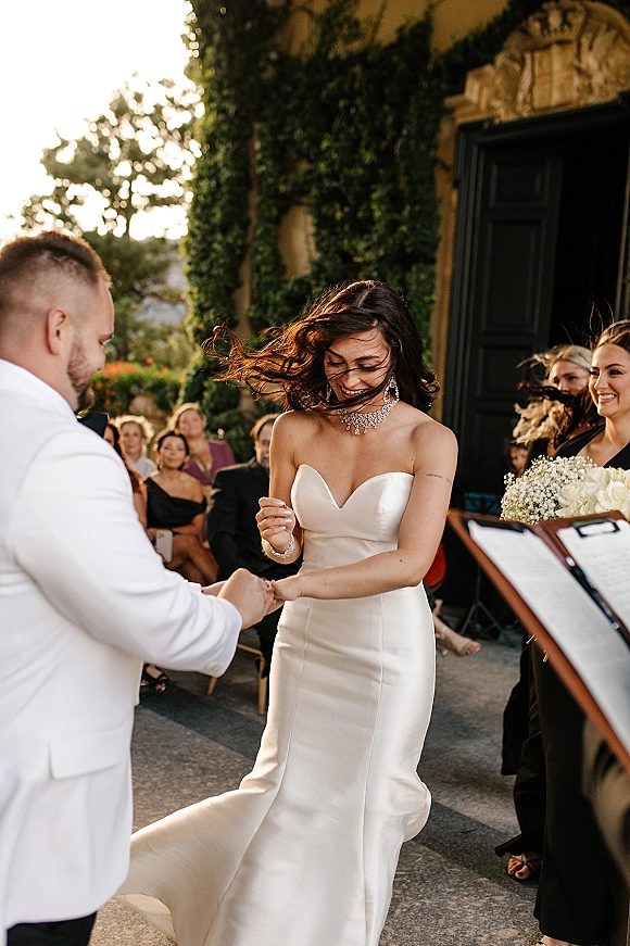Ceremony moment as bride puts on wedding ring, laughing beside groom in white suit at ivy-covered outdoor venue entrance in daylight