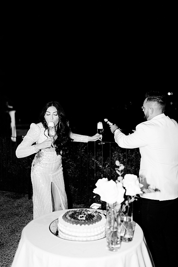 Wedding cake cutting as bride in a sequin gown and groom in a suit slice cake beside champagne flutes on an outdoor patio at night