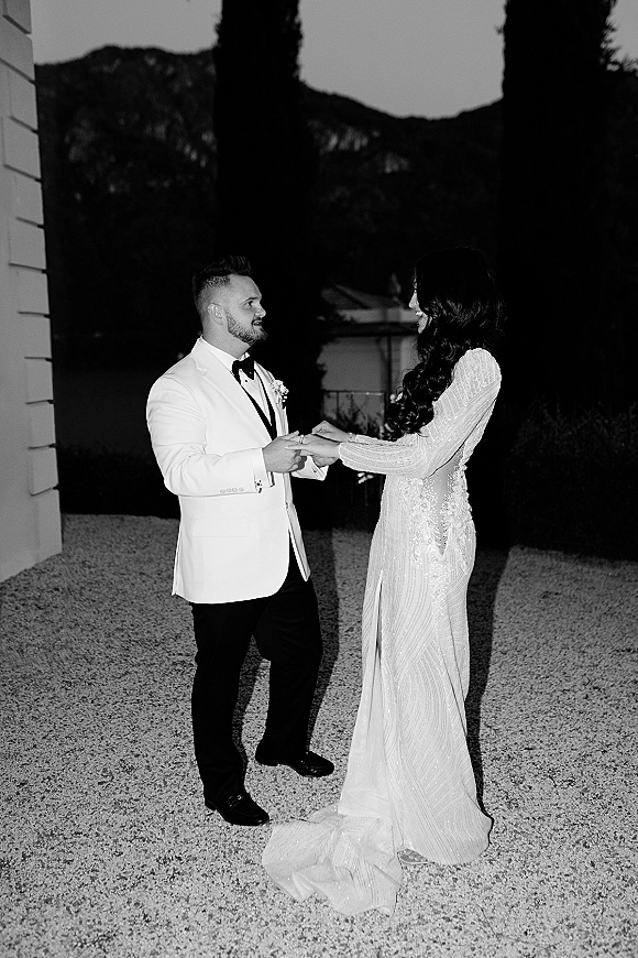 First dance moment as bride in beaded long-sleeve dress with train holds groom in white tuxedo jacket in a gravel courtyard at night