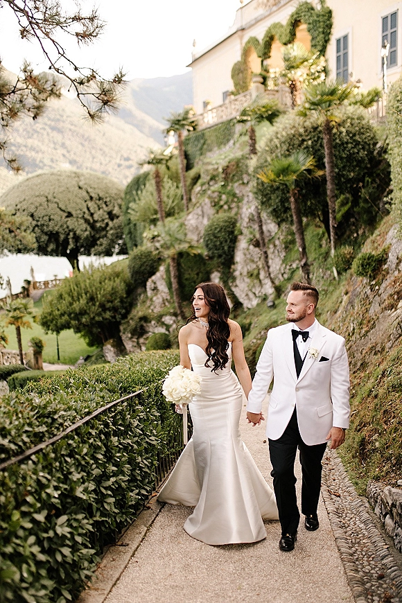 Couple portrait of bride and groom walking hand in hand, her white rose bouquet and long train on a garden path with palm trees and villa backdrop