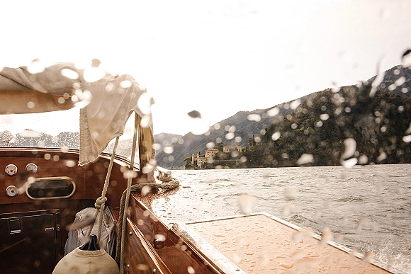 Boat getaway on a wedding boat ride in a vintage wooden speedboat with canopy and rope, crossing a mountain lake past hillside buildings