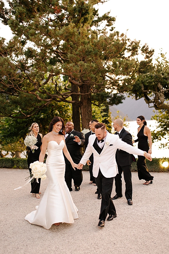 Wedding recessional as bride and groom walking hand in hand with bridal party on a gravel path by a lake and trees under open sky