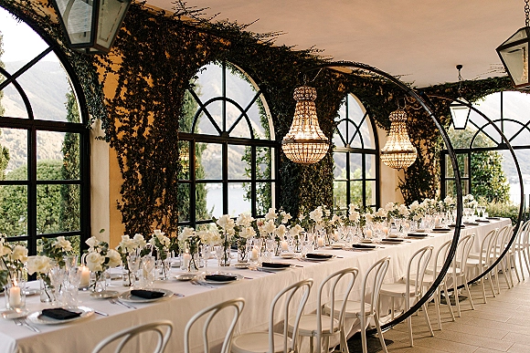 Reception tablescape with a long banquet table wedding setup, white florals in glass bud vases and candles under crystal chandeliers on an ivy terrace