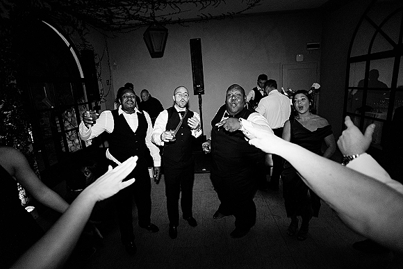 Wedding dance floor packed with wedding party dancing in suits and vests, guests raising cocktail glasses in an indoor room with vines and arches