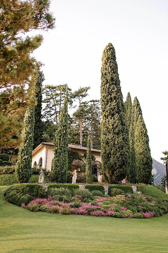 Wedding venue exterior of a villa wedding venue with terracotta roof tiles, arched windows, and stone statues framed by cypress trees and mountains
