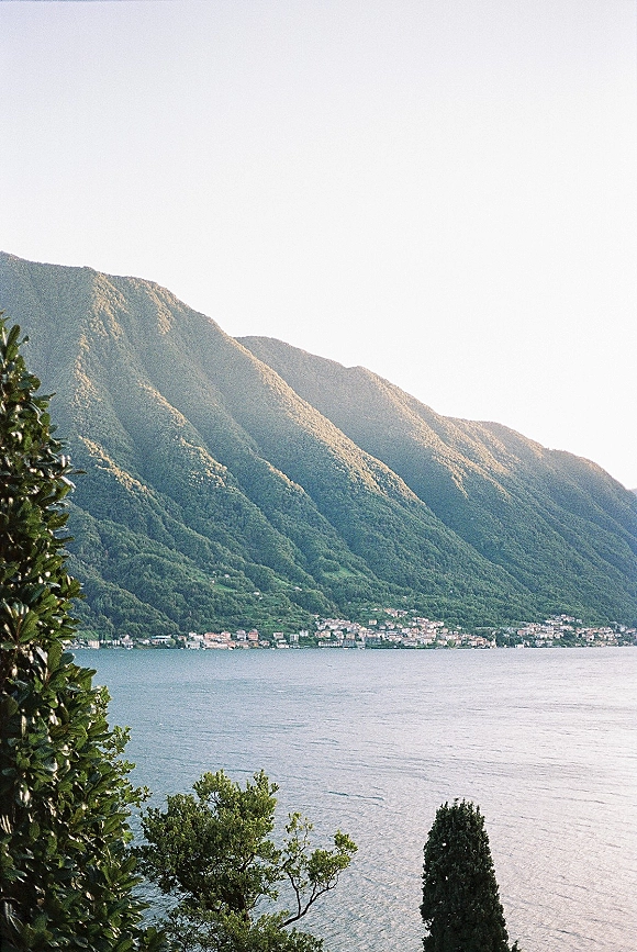 Lake mountain view with calm lake water and trees in the foreground, mountains rising beyond a shoreline town under open sky