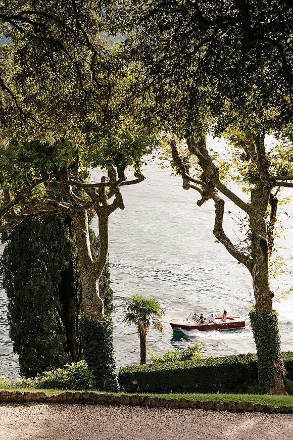 Wedding boat exit as newlyweds walk toward a wooden speedboat on a lake, passing manicured hedges, stone edging, and palms