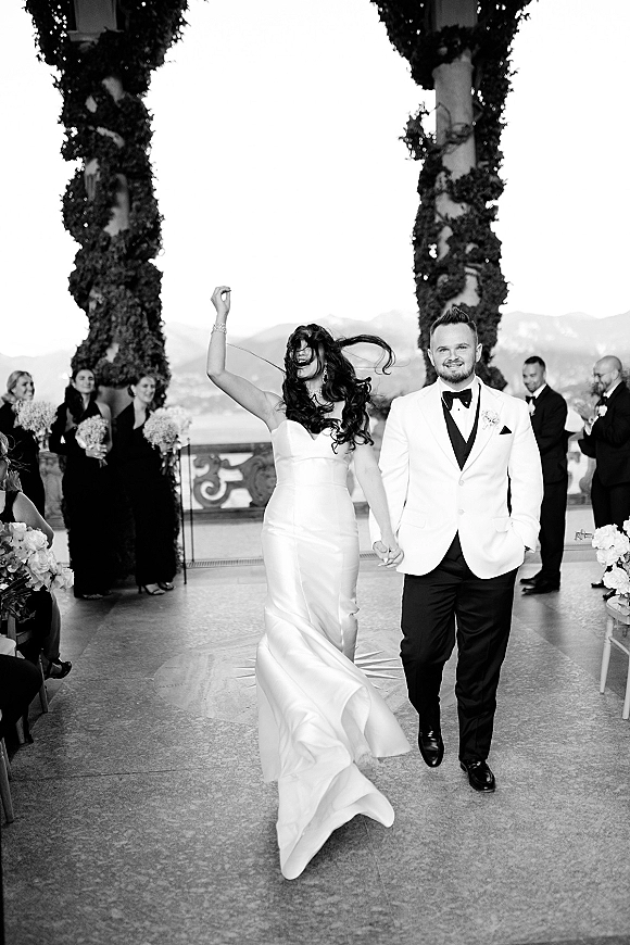 Wedding recessional as bride and groom walk the aisle hand in hand, bride’s train flowing past guests on a mountain lake terrace with columns