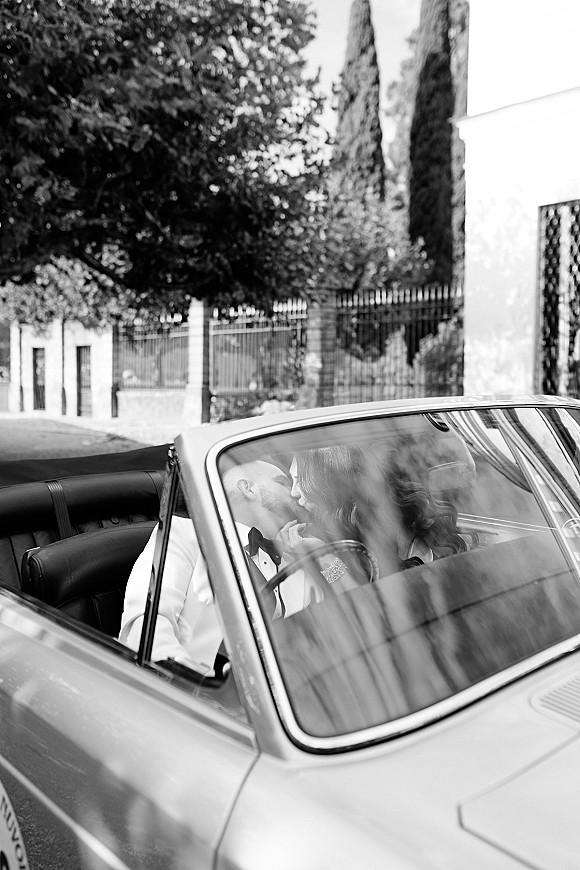 Wedding kiss as newlyweds in car lean together in a classic convertible, bride in statement earrings, street with trees and iron fence behind