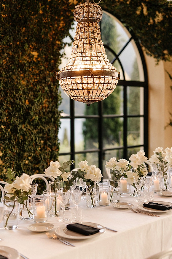 Reception tablescape with a white rose centerpiece, bud vases and floating candles, black napkins, set beneath a crystal chandelier by an arched window ivy wall