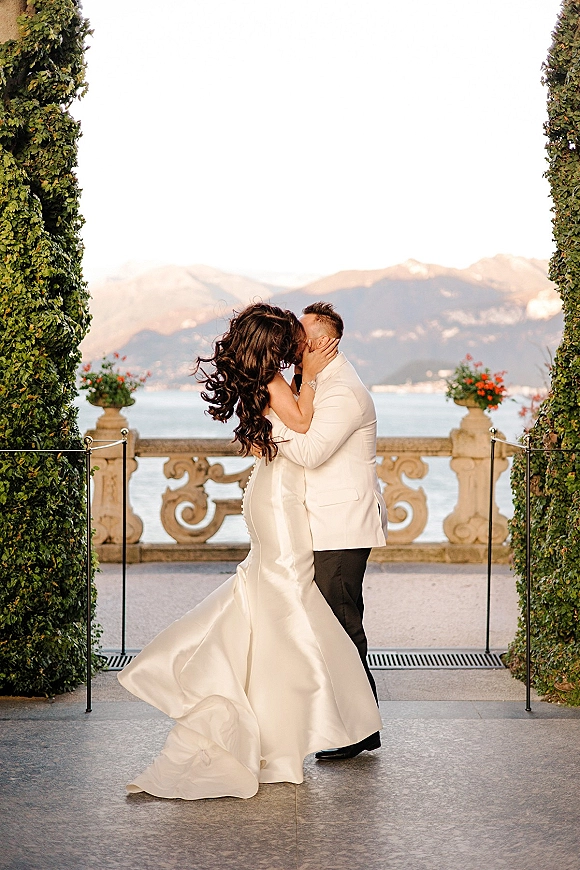 Wedding kiss portrait of bride and groom kissing, bride holding his face on a stone terrace by a lake with mountains behind
