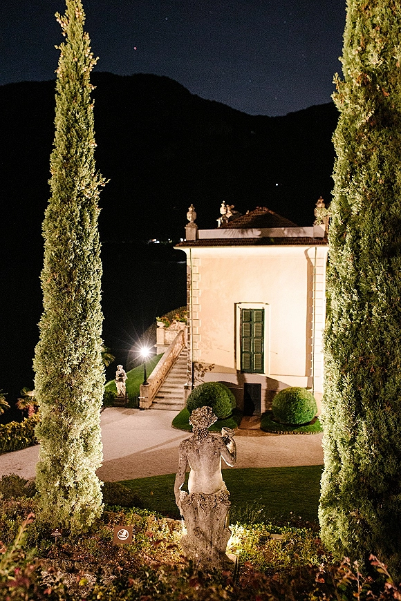 Wedding venue exterior at a villa wedding venue with warm outdoor lights, stone statue, and cypress trees under a night sky with mountains