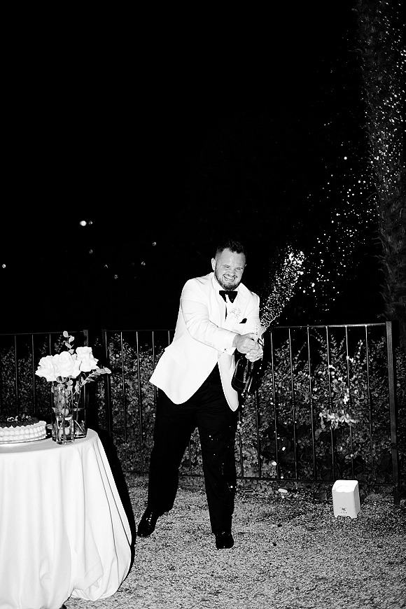 Champagne pop moment as groom in a white dinner jacket pops a bottle, spraying bubbly beside a cake table under the night sky