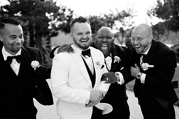 Groomsmen portrait with groom in a white dinner jacket laughing arm in arm with tuxedos and boutonnieres on a tree-lined lawn walkway