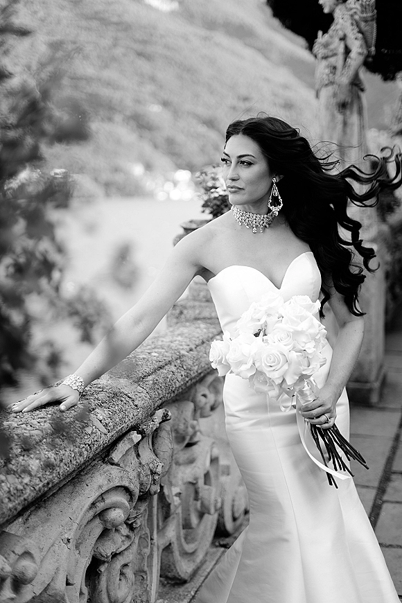 Bridal portrait of a bride holding a white rose bouquet, leaning on a stone balustrade on an outdoor terrace with greenery behind