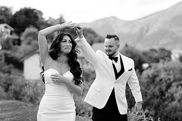 Wedding couple portrait in black and white as the groom spins the laughing bride in a strapless gown, with mountains and trees behind