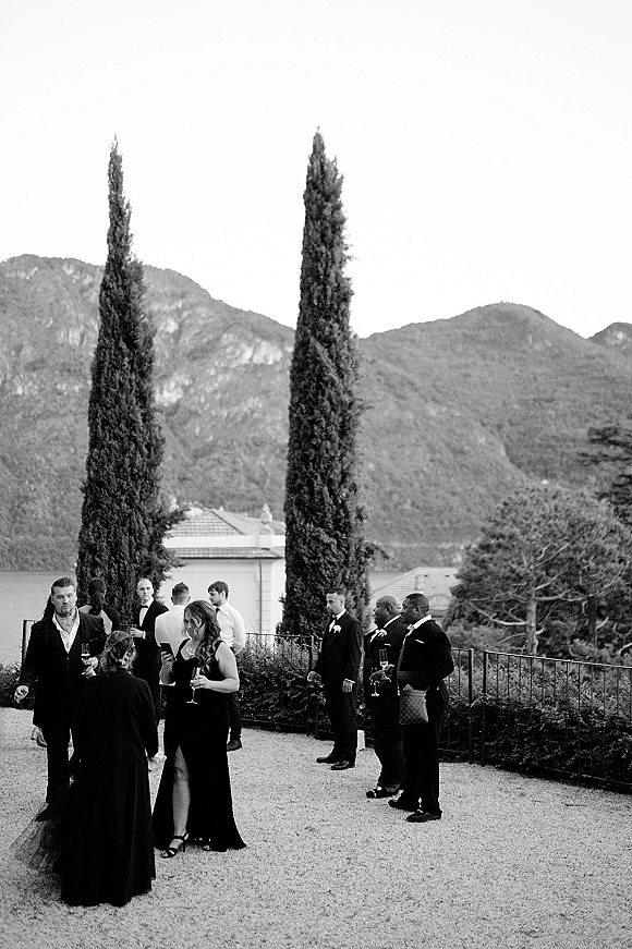 Wedding cocktail hour guests mingling in formalwear with champagne flutes on a villa terrace overlooking mountains and a lake