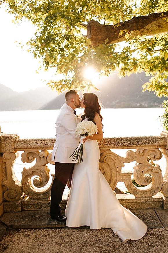 Wedding kiss portrait of bride and groom kissing by a stone balustrade, bride holding a white bouquet with a veil, lake and mountains behind