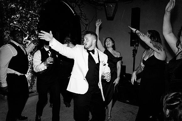 Wedding dance party with groom dancing reception in a white dinner jacket and bow tie, holding a cocktail glass on an ivy-lined patio at night