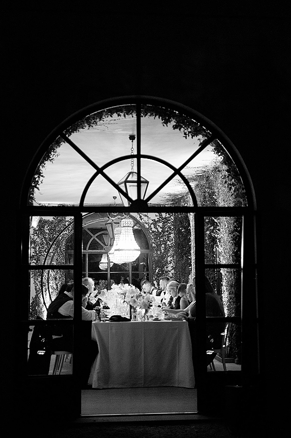 Reception dinner with a wedding guest dinner table on a long banquet table, floral centerpieces and candles beneath a chandelier in an ivy courtyard