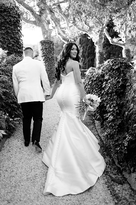 Couple portrait of bride and groom walking away holding hands on a garden path, bride looking back in a strapless dress with bouquet