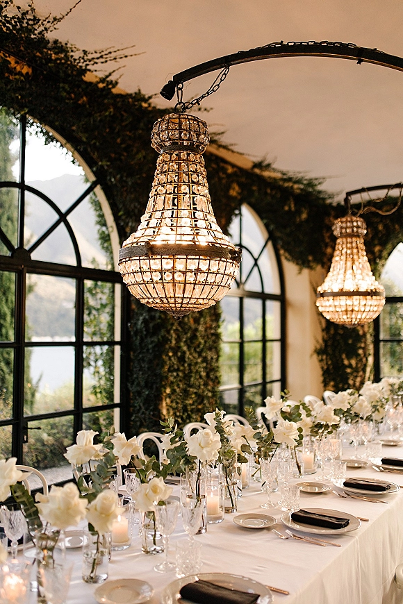 Reception tablescape with wedding table setting of white florals and candles under crystal chandeliers in a greenhouse venue with arched windows